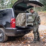 Person in camouflage gear loading a camouflage-patterned case into the back of a black SUV in a forest setting.